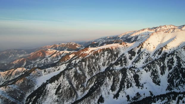 Epic red-orange sunset in high snowy mountains. There are tall coniferous trees, there is snow on the hills. High peaks. The sun's rays fall on the clouds and fog that is lower down the gorge. Almaty