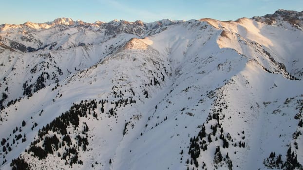 Epic red-orange sunset in high snowy mountains. There are tall coniferous trees, there is snow on the hills. High peaks. The sun's rays fall on the clouds and fog that is lower down the gorge. Almaty