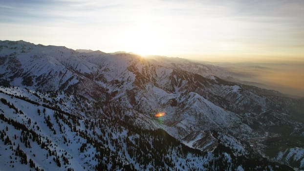 Epic red-orange sunset in high snowy mountains. There are tall coniferous trees, there is snow on the hills. High peaks. The sun's rays fall on the clouds and fog that is lower down the gorge. Almaty