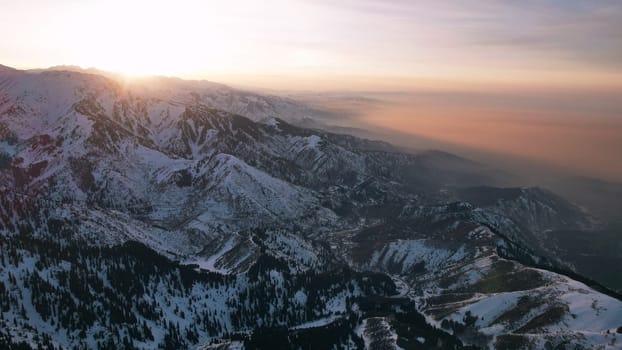 Epic red-orange sunset in high snowy mountains. There are tall coniferous trees, there is snow on the hills. High peaks. The sun's rays fall on the clouds and fog that is lower down the gorge. Almaty