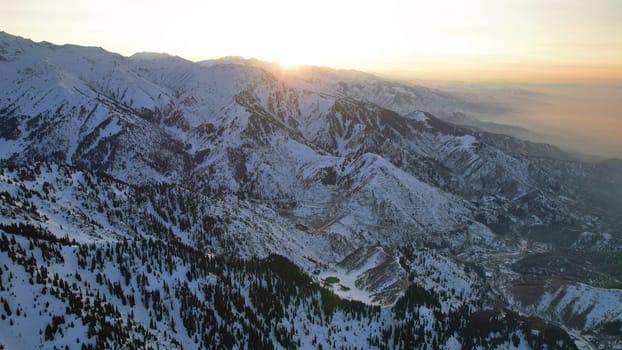 Epic red-orange sunset in high snowy mountains. There are tall coniferous trees, there is snow on the hills. High peaks. The sun's rays fall on the clouds and fog that is lower down the gorge. Almaty