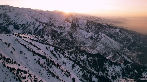 Epic red-orange sunset in high snowy mountains. There are tall coniferous trees, there is snow on the hills. High peaks. The sun's rays fall on the clouds and fog that is lower down the gorge. Almaty