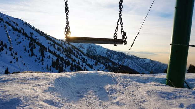 Swing in winter mountains with sunset view. The sun goes behind the snowy mountains. Coniferous trees are visible at the bottom of the gorge, there is a forest there. Orange light on the clouds
