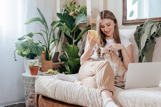 Freelance woman with dog and laptop, texting at mobile phone and working from home office. Happy girl sitting on couch in living room with plants. Distance learning online education and work.