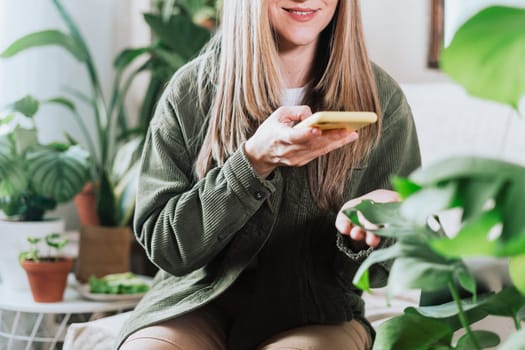 Cropped woman sitting on the couch and taking photo of a lot of plants. Cozy home and urban jungle concept. Woman with mobile cellphone and plants.