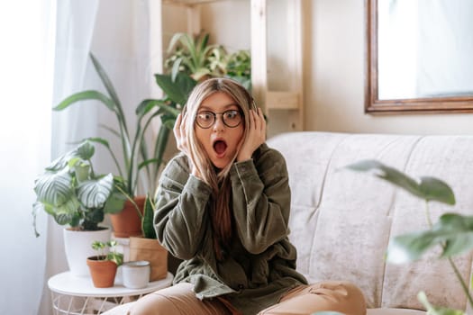 Shocked woman with open mouth in glasses listening music in headphones and relax at home. Frustrated girl sitting on couch in living room with plants. Urban jungle concept