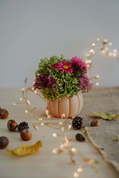 Autumn bouquet in a decorative planter in the form of a pumpkin. Autumn composition. Happy Thanksgiving. Bokeh from a garland. Festive image. Cones and nuts. Vertical photo.