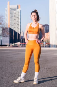 vertical photo of a female runner standing with hands on hips and looking at camera in the city, concept of active lifestyle and urban sport