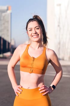 vertical portrait of a female runner smiling happy with hands on hips and looking at camera in the city, concept of active lifestyle and urban sport