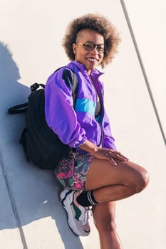 vertical portrait of a smiling african american young woman in sports clothes leaning on an urban wall, concept of active lifestyle and urban sport, copy space for text