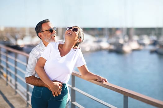 Summer Love Story. Romantic Senior Couple Embracing Expressing Happiness Standing At Marina Pier Outside, Smiling And Laughing In Joy On Summer Day At Seaside. Sea Holidays In Older Age. Free Space