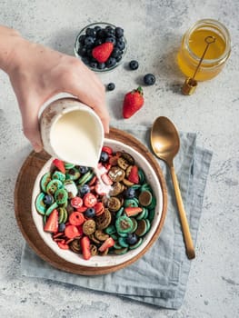 Trendy food - pancake cereal. Milk pouring on heap of multicolor mini cereal pancakes in bowl. Tiny cereal pancakes with berries in craft plate over gray cement background. Vertical. Top view,flat lay