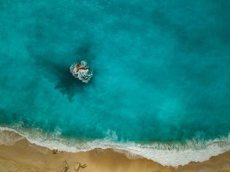 Drone shot of beautiful crystal clear turquoise and blue sea water, surrounding a rock and waves reaching sandy beach. 