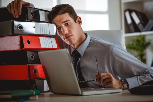 Frustrated young handsome man looking exhausted and leaning at the binders, while sitting at his working place