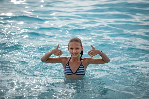 Cute little girl enjoing in the sea water on the beach, showing thumbs up and smiling looking at camera.