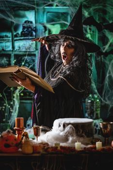Witch with awfully face in creepy surroundings and smoky green background reading recipe of magic drink sends evil. Halloween concept.