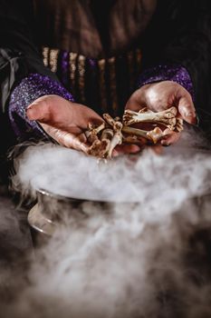 Close-up of a witches hands holding bones above boiling water for a cooking poison soup.