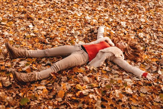 Cute smiling young woman lying down on the fall leaves and enjoying in the nature in autumn.