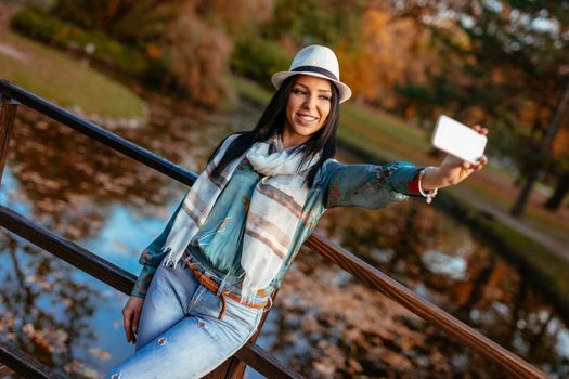 Modern cute girl enjoying to take a selfie on the bridge in autumn city park.  