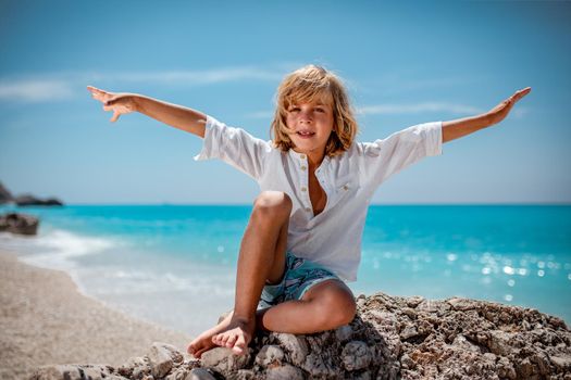 Cute boy is sitting on the sea rock on the beach and smiling looking at camera. His arms are wide open.  
