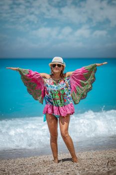 Beautiful young woman in colorful dress with open arms, and summer hat on her head, enjoying at sun in the beach.
