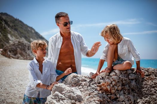 Beautiful little boys enjoying with their father on the beach. One boy is sitting on the rock, and the other boy and a father looking at him.