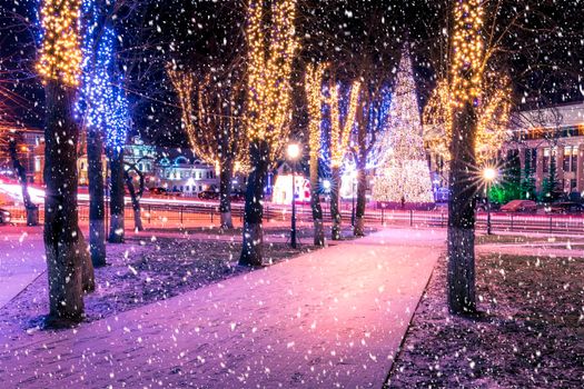 Snowfall in a winter park at night with christmas decorations, lights, pavement covered with snow and trees with garlands.