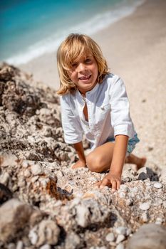 Cute boy is climbing up on the sea rock on the beach, smiling and looking at camera.