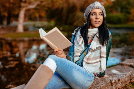 Beautiful young pensive woman enjoying in autumn colors near the little lake in the city park looking away and holding a book in her hand.  