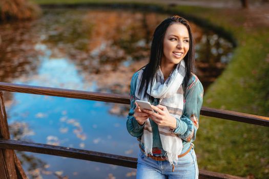 Young happy woman on a wooden bridge over the little lake in the park enjoying in beautiful autumn golden day with a smartphone.