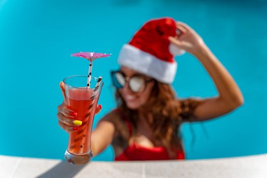 Young beautiful woman near the swimming pool in Santa Claus hat celebrating New Year and Christmas in hot country with glass of cocktail.
