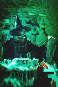 Witch with awfully face in creepy smoky green surroundings full of cobweb is reading recipe of magic drink. Halloween concept.