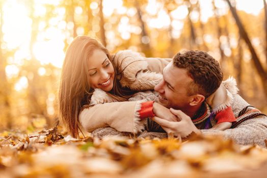 Beautiful smiling couple enjoying in sunny forest in autumn colors. They are lying down on the fall leaves and having fun.