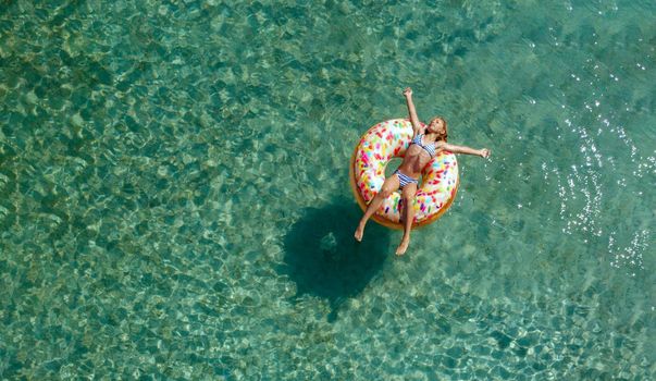 Aerial view of little girl swimming on the inflatable big donut in the transparent turquoise sea. Top view from drone.