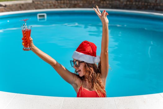 Young beautiful woman near the swimming pool in Santa Claus hat celebrating New Year and Christmas in hot country with glass of cocktail.
