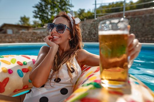 Beautiful young woman in swimming pool swims on inflatable ring donut and has fun with glass of beer on vacation.