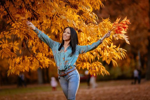 Cheerful young beautiful woman going alone in the park and holding a branch of a fallen leaves in golden sunny autumn.