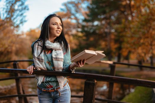 Young happy woman on a wooden bridge over the little lake in the park enjoying in beautiful autumn golden day with a book.