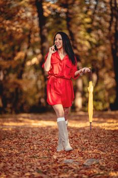 Happy young woman in red dress talking on smartphone walking in autumn sunny park, holding folded yellow umbrella.