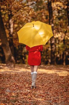 Young beautiful girl in red dress going alone under yellow umbrella in the park in golden autumn. Rear view.