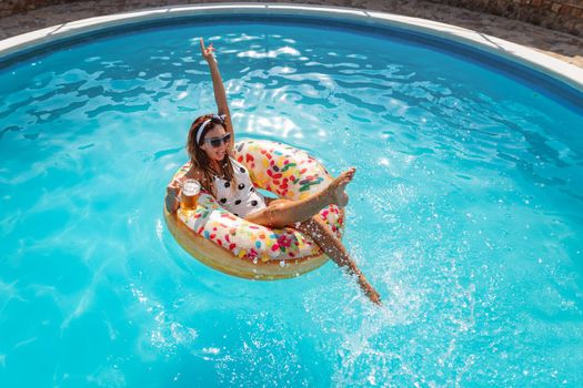 Beautiful young woman in swimming pool swims on inflatable ring donut and has fun with glass of beer on vacation.