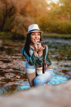 Happy woman photographer holding professional digital camera and taking photo near the lake in autumn park.  