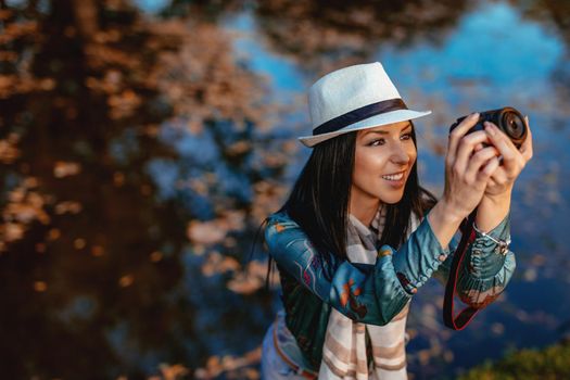 Happy woman photographer holding professional digital camera and taking photo near the lake in autumn park.  