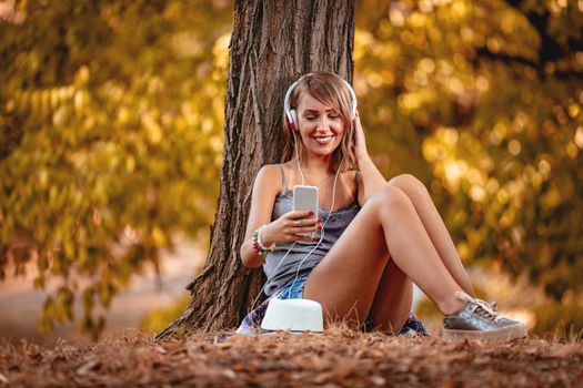 Cute smiling young woman sitting on the fall leaves in early autumn sunny day and listening music.