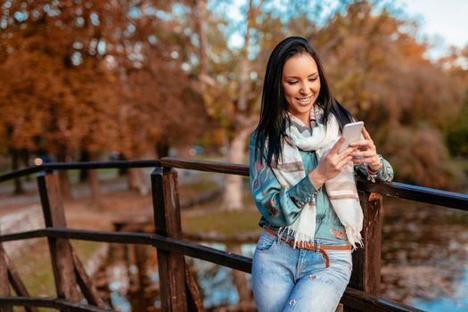 Happy young smiling woman reading message with pleasant news on smartphone in autumn nature.