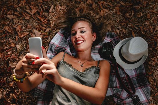 Top view of a cute girl lying down on the fall leaves and enjoying taking selfie in the nature in early autumn sunny day.