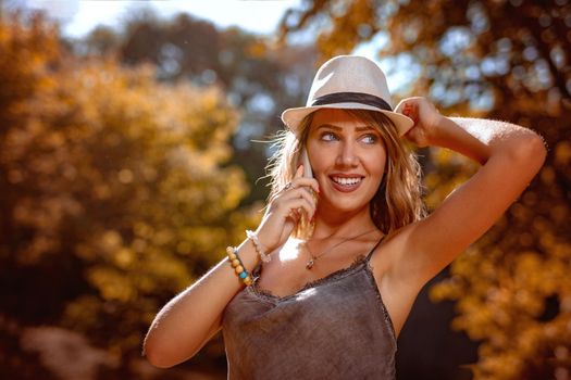 Beautiful smiling young woman with summer hat enjoying in the nature in autumn and talking on smartphone.