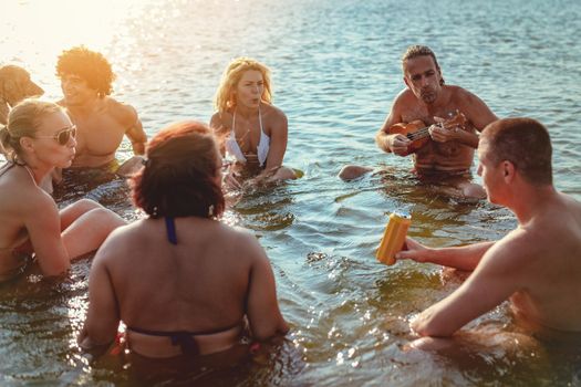 Happy young people having a great time together at the beach. They are sitting in the water and man is playing ukulele and singing. Sunset over water.