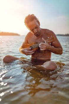 Happy young musician has a great time at the beach. He is sitting in the water, playing ukulele and singing. Sunset over water.