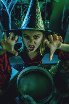 Top view of young witch with awfully face looking at camera and tells evil words above cauldron.